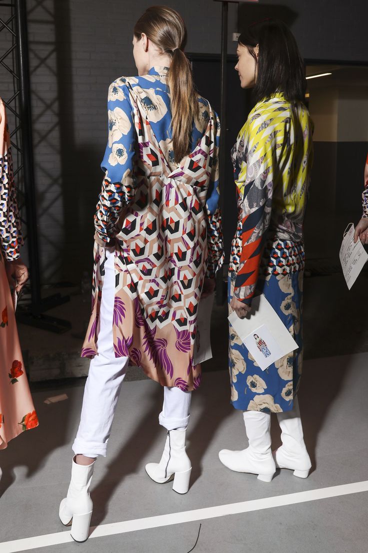 wo models stand backstage at a fashion show. they are both wearing longsleeved patterned dresses with geometric and floral prints, white pants, and white ankle boots. one model is wearing a predominantly blue, yellow, and white dress, while the other is wearing a dress with more pink and beige tones.  both dresses are loosefitting and fall to midcalf. the background is somewhat blurred, showing a metal structure and a glimpse of another model in a different outfit.  the atmosphere is suggestive of a postshow moment.