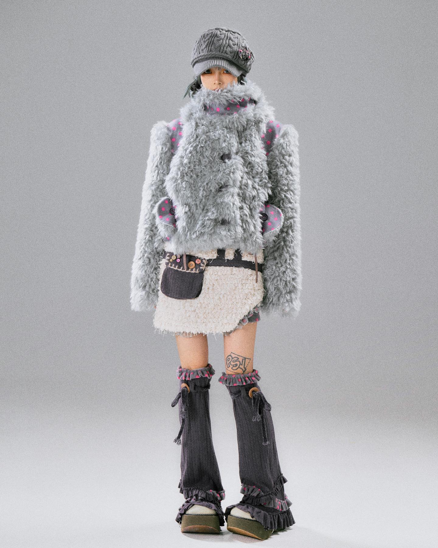 the image showcases a fashion model wearing a unique and stylish winter outfit. the centerpiece is a fluffy, light grey oversized jacket with subtle polka dot detailing on the sleeves.  underneath, she wears a creamcolored, textured miniskirt with a high waist. the skirt has a slightly layered and ruffled appearance.  the model also sports dark grey leg warmers with frilly hems, and platform shoes that are dark in color and have a similar ruffled detail around the ankles. she accessorizes with a grey newsboy cap and a small, darkcolored pouch bag at her waist.  the overall style is avantgarde, combining textures and whimsical elements for a striking look. the backdrop is a plain, light grey studio background, drawing focus entirely to the model and her outfit.