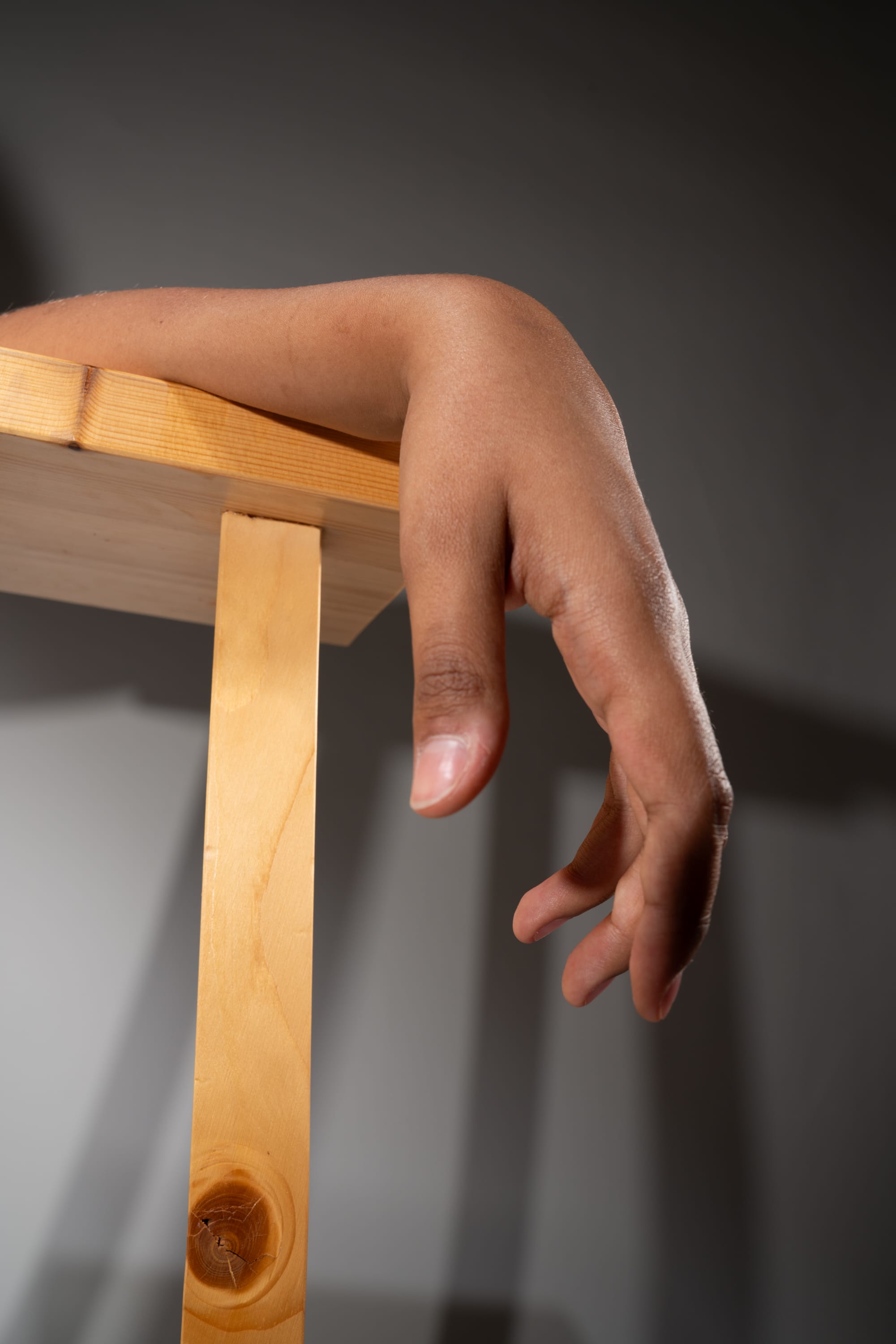 a closeup, worm's eye view of a person's arm resting on a light brown wooden table. the hand is relaxed, with fingers slightly curled. the image uses soft lighting, creating subtle shadows on the table and wall in the background.