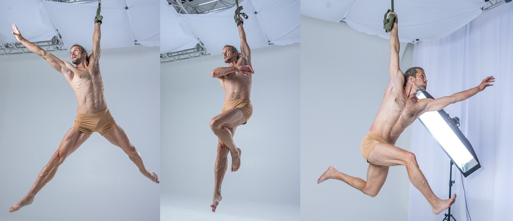 a muscular man wearing tan shorts is suspended midair in a white studio setting. he is performing a variety of gymnastic poses, demonstrating flexibility and strength.  the man is shirtless and his muscles are clearly visible. he is suspended by a set of straps that appear to be attached to his hands.  there is a white backdrop in the background.  the man's face is visible, but there is no expression on it.