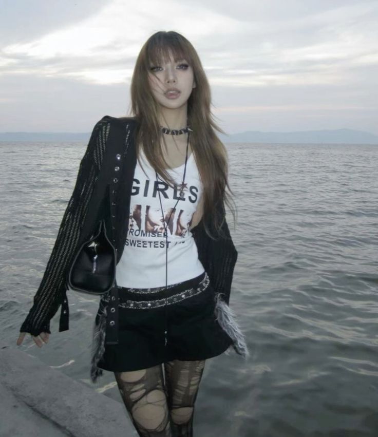 a young woman with long brown hair is standing by the ocean. she is wearing a black cardigan, a white graphic tshirt with the word "girls" on it, a black mini skirt, ripped fishnet stockings and a black handbag. she has on a black choker necklace. the overall style is grunge/goth. the background is a calm ocean with a slightly cloudy sky.