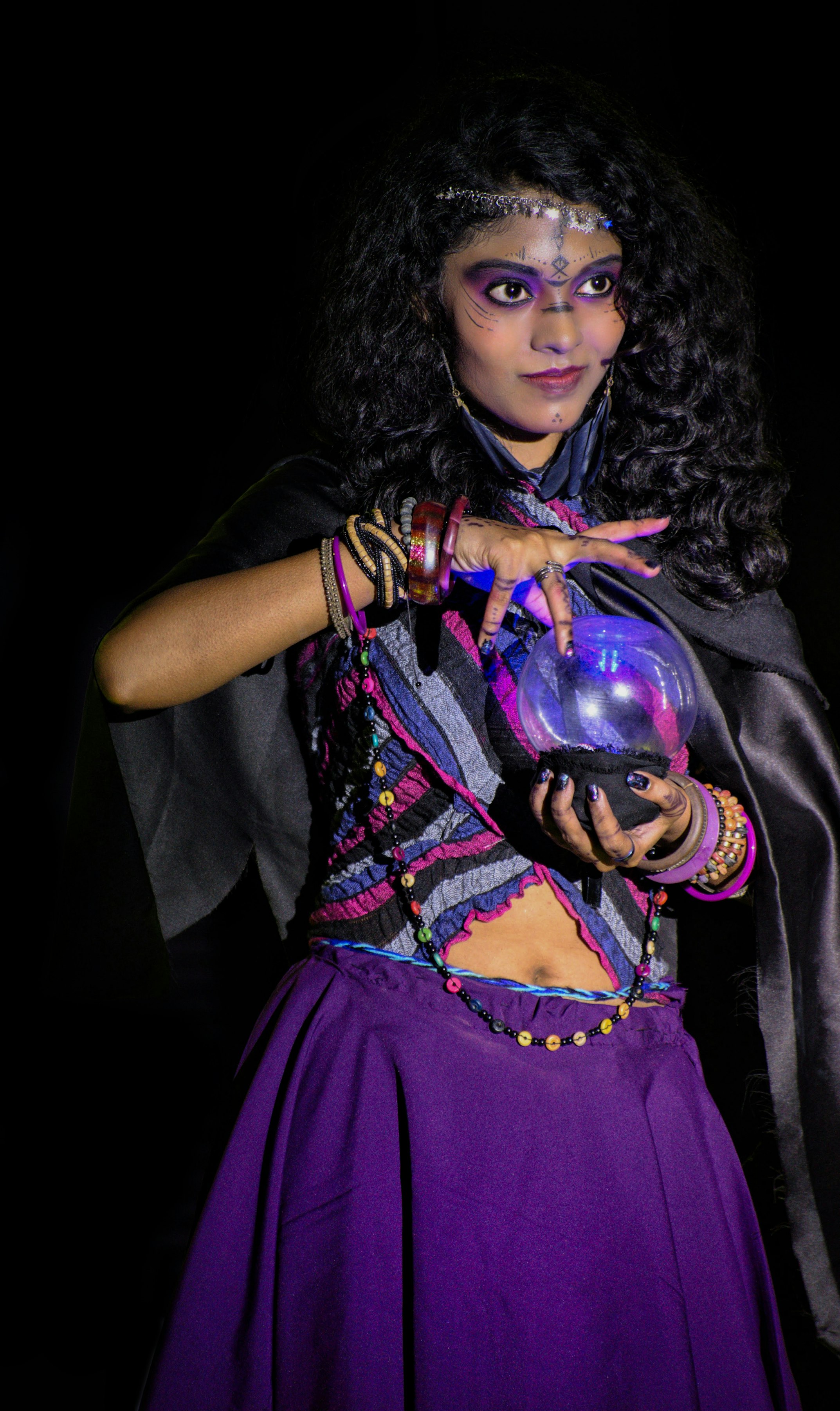 the image depicts a young woman dressed in a mystical costume, possibly for cosplay or a theatrical performance. she is portraying a fortune teller or similar character.  she's wearing a purple dress with layered bohemianstyle clothing, adorned with various necklaces and bracelets. her makeup includes face paint, enhancing the mystical aesthetic.  she holds a crystal ball, which is subtly lit from within, creating a glowing effect. her dark, curly hair frames her face, and she appears to be gazing intently into the crystal ball. the background is dark, enhancing the mysterious ambiance of the scene. the overall mood is one of intrigue and magic.