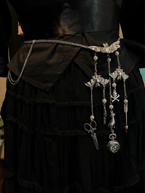 a closeup shot showcases a woman's waist adorned with a gothicinspired fashion ensemble. she is wearing a black dress with a tiered skirt and a wide black belt cinching her waist. a silver chain belt, embellished with batshaped pendants, pearls, and various charms such as scissors, a hand, a watch, a skull, and an arrow, dangles from the belt. the overall style evokes a victorian or gothic aesthetic, highlighting the intricate details of the dress and accessories.