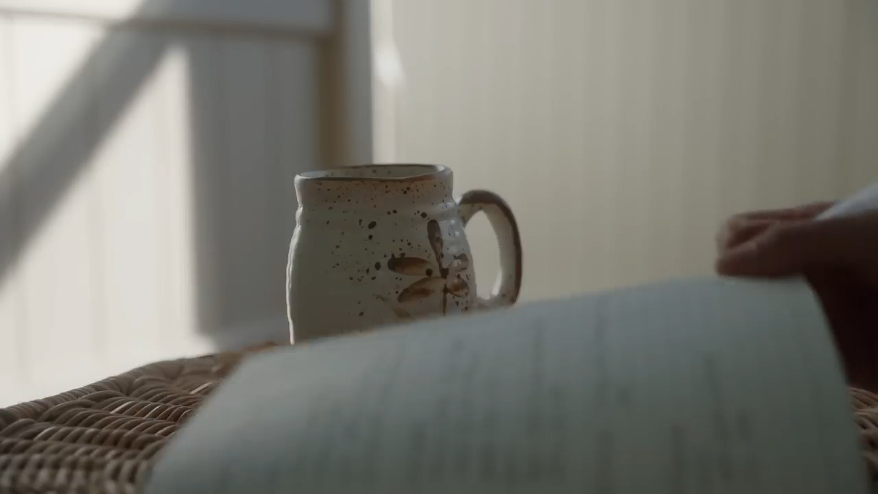 a closeup shot of a book open on a wicker surface.  a white mug with a brown design and a partially visible hand holding the book are slightly out of focus.  the background is out of focus and shows the interior of a bright sunlit room.