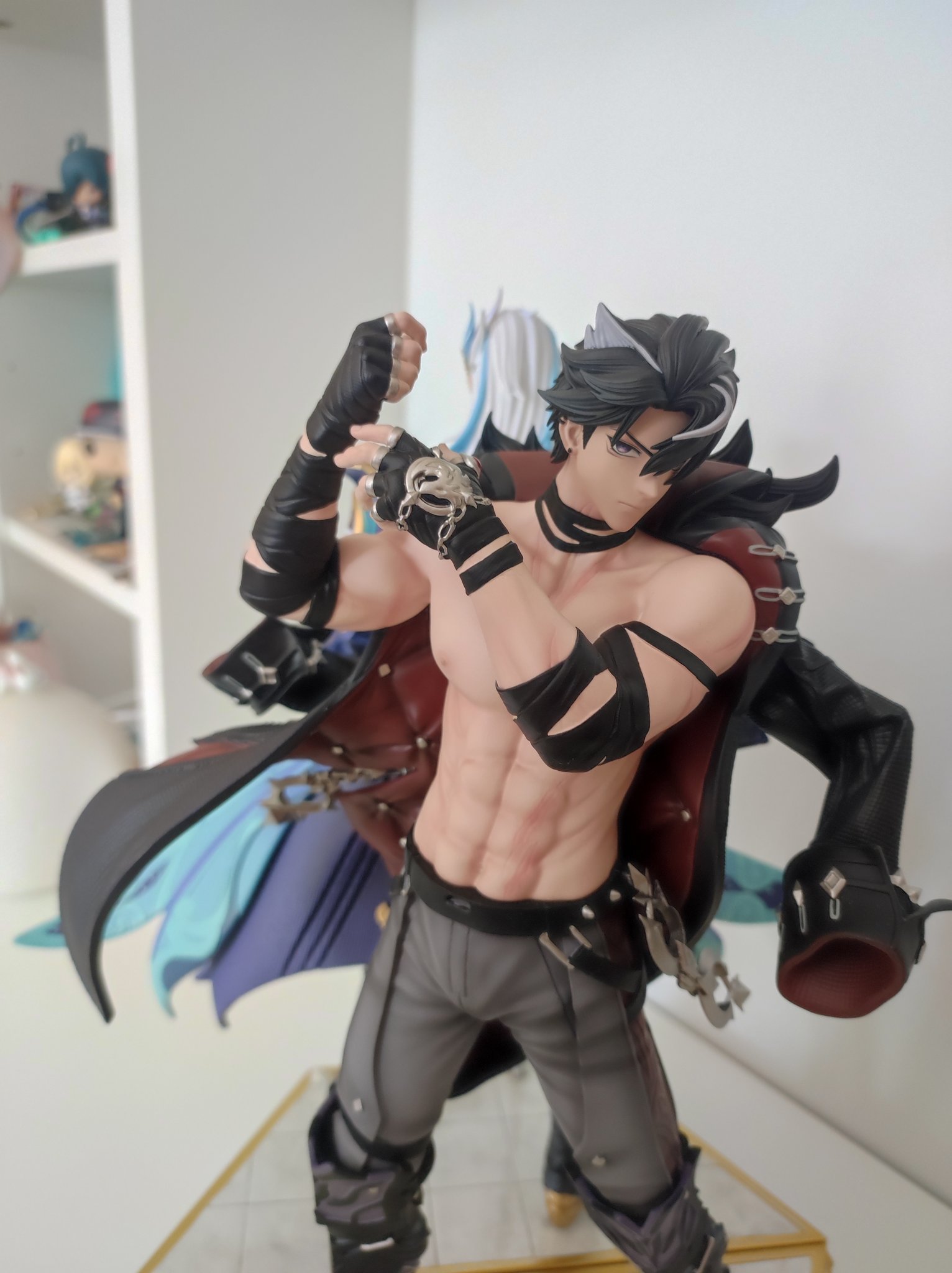 a figurine of a shirtless anime character with black gloves, black and red clothes, a silver chain, and grey pants. he's posing and looking toward the right side of the image.  the figurine is standing on a platform, and there is a blurry image of another figure in the background.