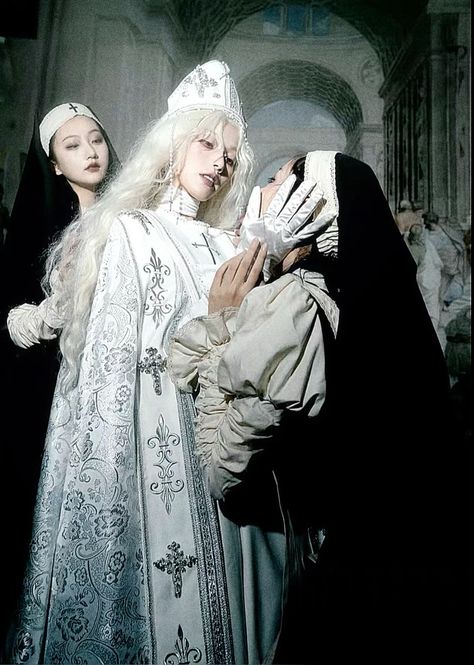 three women pose dramatically in a dimly lit church. they are all dressed in elaborate, historically inspired costumes resembling nun's habits. one woman in a tall white pointed hat and ornate robe stands in the center, framed by the other two.  the lighting and poses suggest a theatrical or storytelling element.