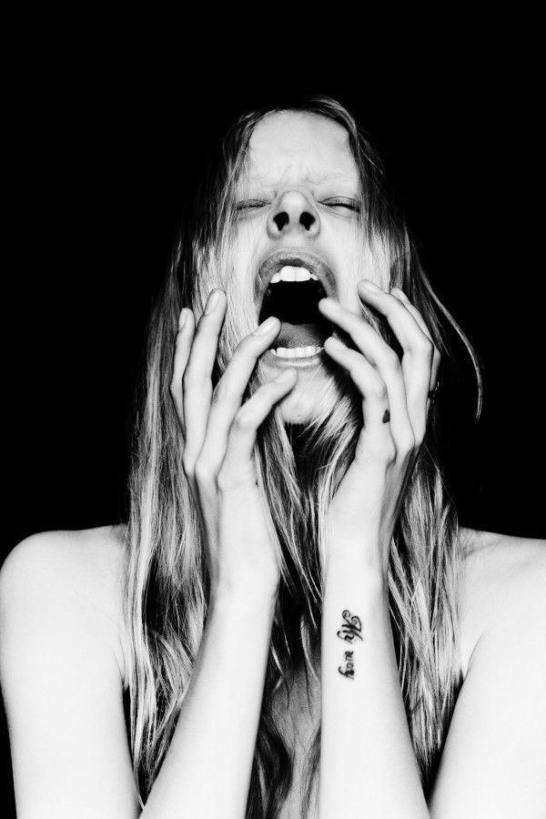 a black and white portrait of a young woman screaming with her mouth wide open. her hair is long and flowing, and she has a tattoo on her wrist. the image is taken against a dark background.  the image is dramatic and intense, conveying the woman's raw emotion.