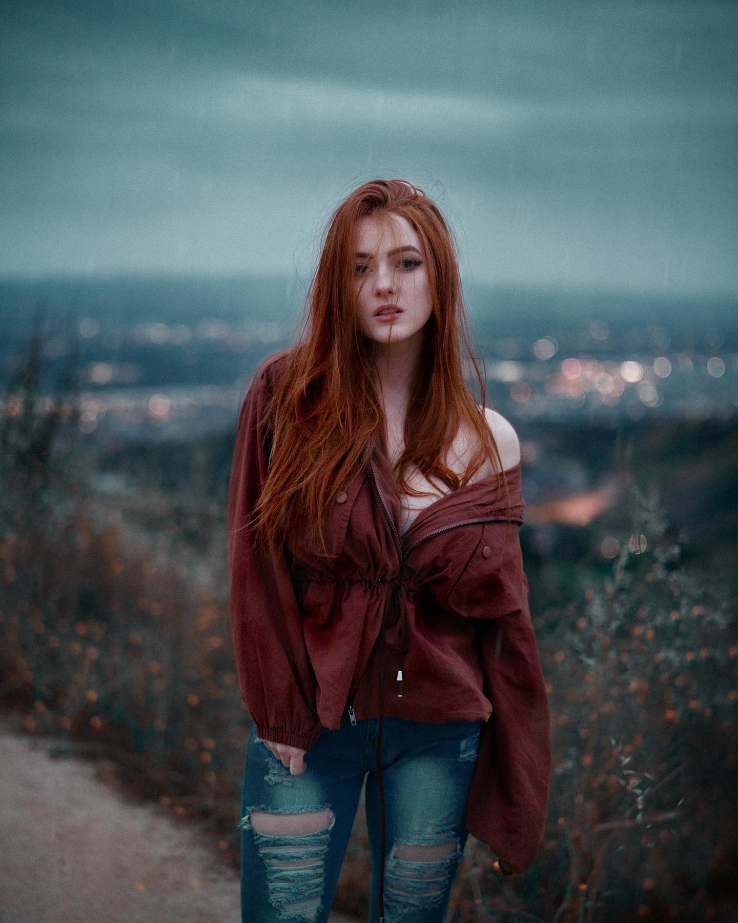 he image shows a young woman with long red hair standing on a dirt path. she is wearing a maroon jacket and ripped jeans. the background is a blurred view of a hillside with a town in the distance. the sky is overcast and dusk is approaching, giving the image a moody atmosphere. the lighting is soft and natural.