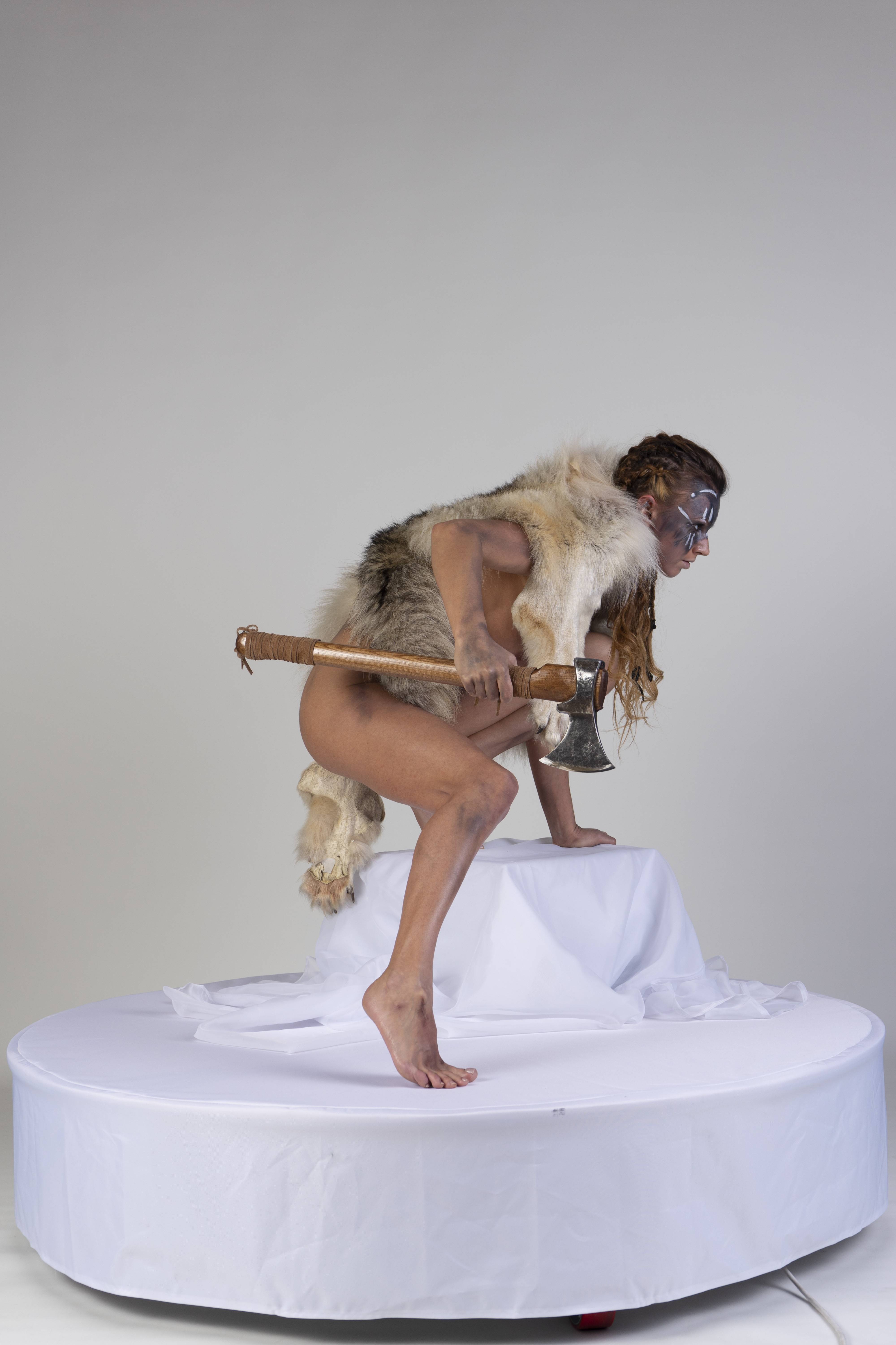 a woman in a viking warrior pose, wearing body paint and a fur loincloth, stands on a white platform with an axe. she is barefoot and appears to be crouching or moving forward. the background is a plain light grey.