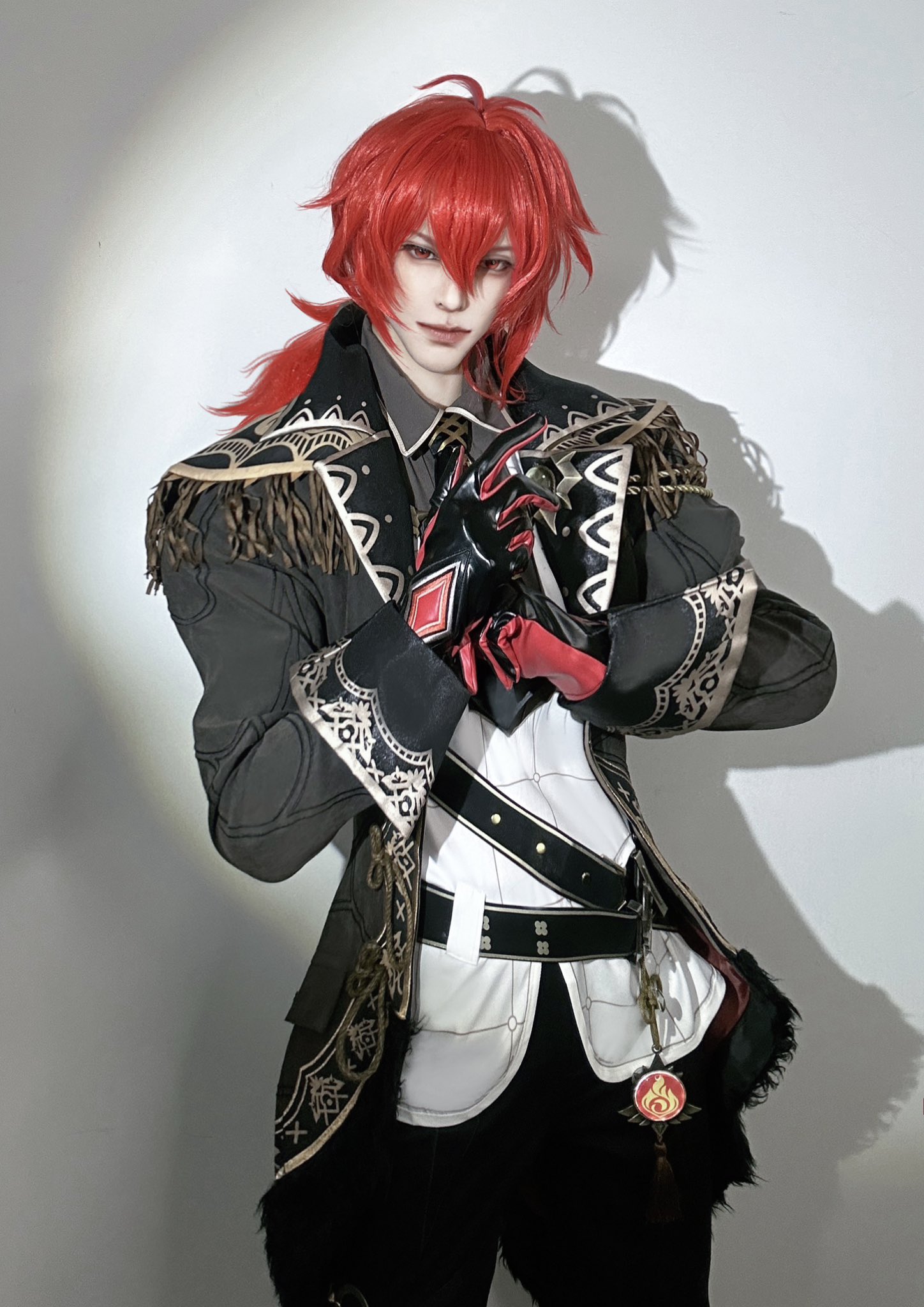 a person is cosplaying a male anime character. he is wearing a black jacket with gold trim and white shirt. he has red hair and is wearing black gloves and pants. he is also wearing a belt and a pendant with a red and gold symbol.  the person's eyes are dark, and their lips are painted red. they are holding a small, gold object in their right hand.  the background is a plain white wall. the photo is taken from a slightly low angle.