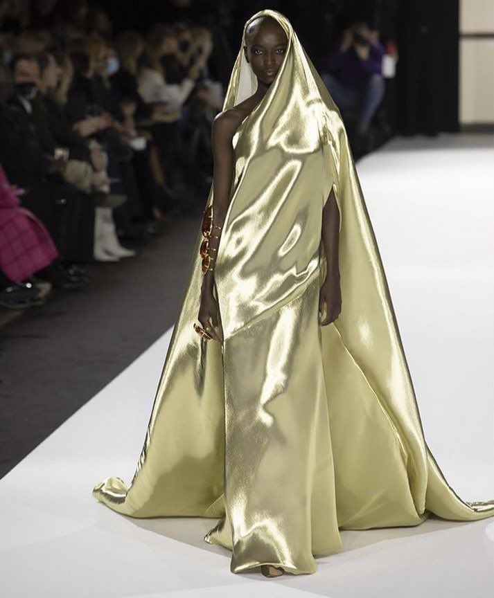 a model walks the runway in a gold satin gown with an attached cape. the gown has a oneshoulder design and the cape is draped over her head. she is wearing jewelry on her arm and hand. an audience watches from the side.