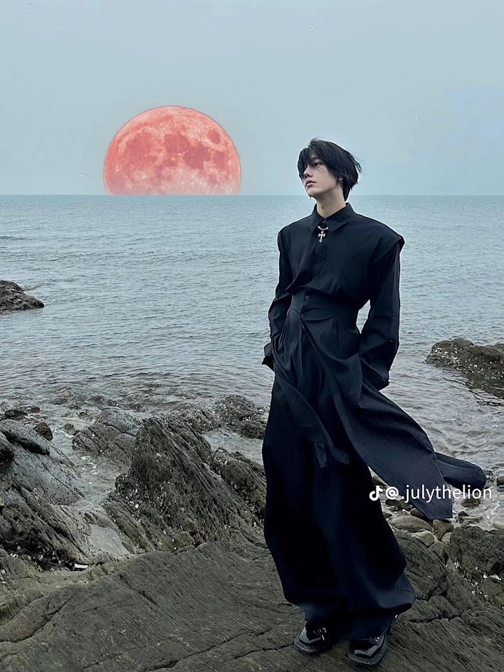 a young man with black hair and a black outfit stands on a rocky shoreline against a backdrop of a red moon and blue ocean, the moody atmosphere creates a cold and dramatic tone.  the individual's black attire contrasts against the gray sky and creates a striking silhouette. the rocky shoreline adds a raw and natural element to the portrait.
