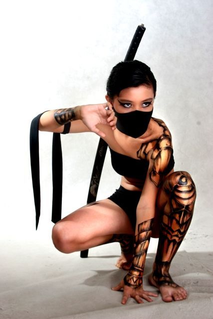 a beautiful woman is posing in a studio with a katana sword. her body is painted with intricate details, and she is wearing a black mask. she looks powerful and ready to fight. the image is welllit and composed.