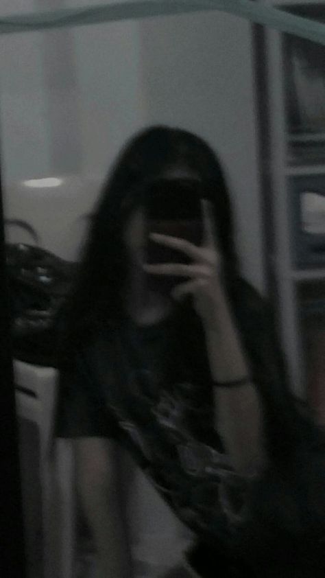 a blurry grayscale selfie of a girl with long black hair taking a picture in a mirror with her phone. the image is dark and the girl's face is obscured by the phone. she is wearing a tshirt and a bracelet.