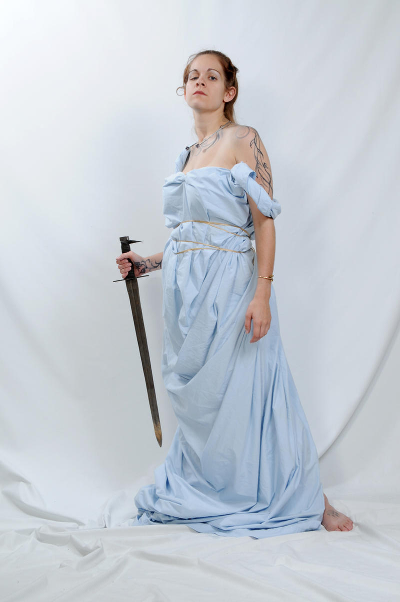 a woman stands in a studio against a white background, dressed in a light blue togalike garment secured with bands around her waist. she is barefoot and holds a sword in her left hand. tattoos are visible on her shoulders and arm. she looks directly at the camera with a serious expression. the toga is draped and flowing, creating a classical or historical aesthetic. the background is a simple white fabric.