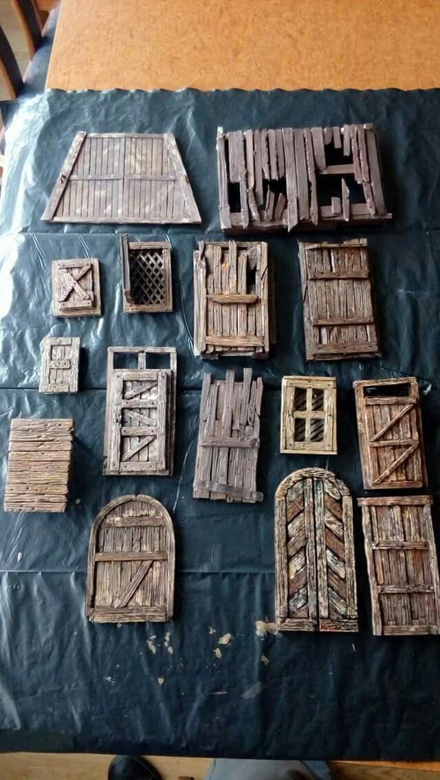the image displays a collection of handcrafted miniature doors and windows, likely made from wood, arranged on a dark, possibly plastic, surface. these appear to be models or props, perhaps for tabletop games such as dungeons and dragons. the doors are of varying designs, some rustic or damaged in appearance, adding a fantasy or medieval aesthetic.