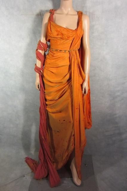 a full shot of a mannequin wearing a long, burnt orange dress styled in an ancient greek or roman fashion.  the dress is draped and cinched at the waist with a belt. it shows signs of wear, such as discoloration and small holes, suggesting it's a costume possibly from a theatrical production or movie.  a reddishorange stole or fabric is also wrapped around one arm and trails to the floor. the background is a neutral, textured gray.