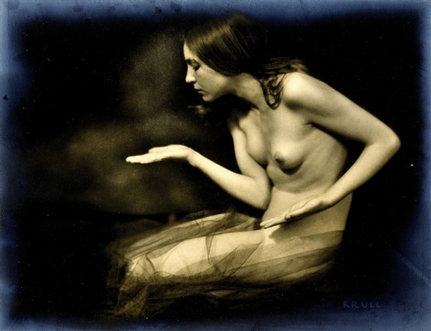 the image is a vintage, sepiatoned photograph of a nude woman. she is seated, partially draped in a sheer, dark fabric that resembles tulle or gauze. her pose is evocative, almost dreamlike, with her hands gently gesturing, as if scattering dust or ashes. her expression is serene and introspective. the background is dark and shadowy, focusing attention on the model’s form. the overall mood is sensual, artistic, and mysterious, reminiscent of vintage nude photography from the early to mid 20th century. the photograph is signed "kr.ull" in the bottom right corner, likely identifying the artist.