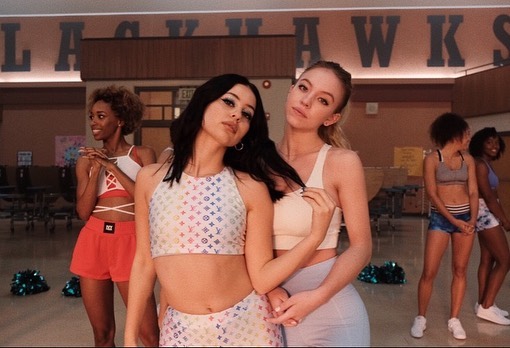 two teenage girls, one blonde and one brunette, are standing close together in a high school gymnasium. they are wearing matching pastelcolored athletic outfits with a louis vuittoninspired pattern on the crop top and leggings. the blonde girl is standing slightly behind and to the side of the brunette girl, her arm around her waist. both girls are smiling, suggesting a friendly relationship. in the background, other cheerleaders are visible, some standing in groups and others in the process of warming up. the overall atmosphere is cheerful and suggestive of a highschool sports scene.