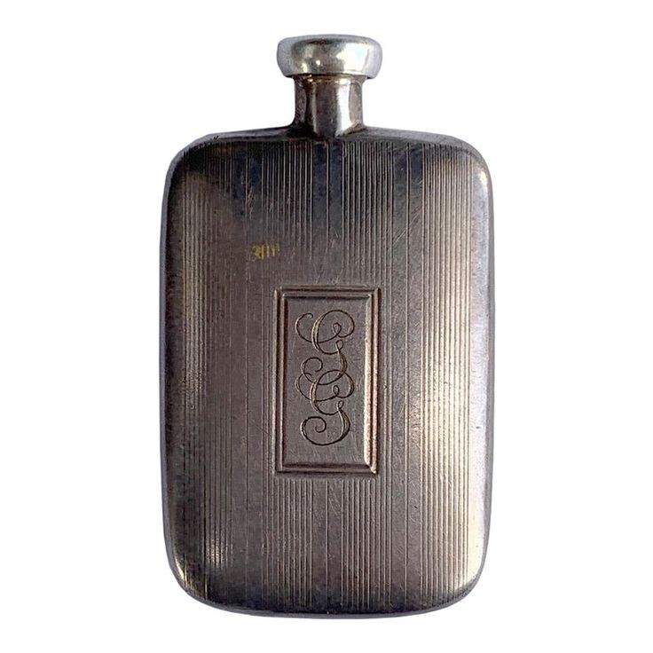 a vintage silver flask with an engraved monogram on the front. the flask is rectangular with rounded edges and a screwtop lid. it has vertical lines on the front and back, with a rectangular frame surrounding the monogram.