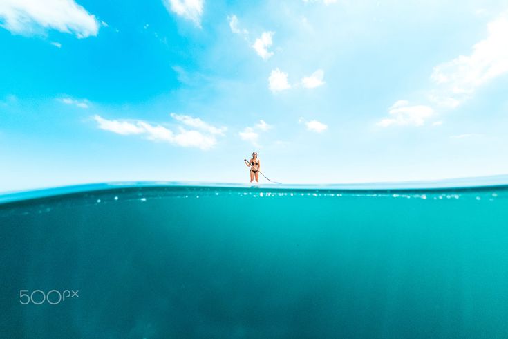 the image is a split shot, with the top half showing a woman paddleboarding on calm, turquoise water under a bright blue sky with fluffy white clouds. the bottom half of the image is underwater, showcasing the same vibrant blue water, hinting at the ocean's depth.