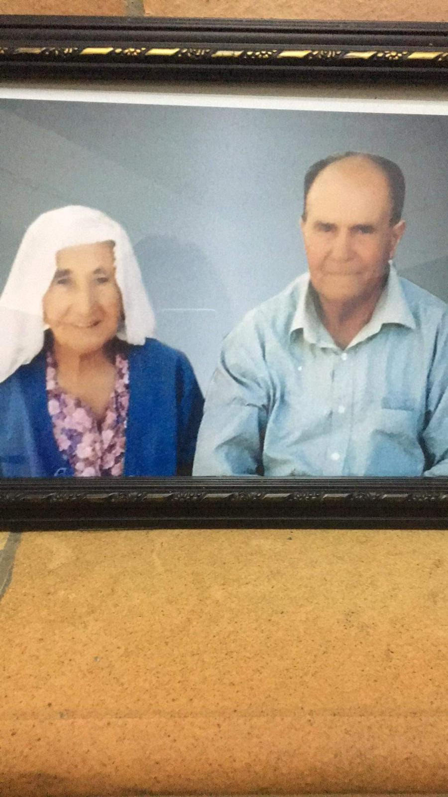 a framed photograph shows an elderly couple. the woman is wearing a white head covering and a blue robe. the man is wearing a light blue shirt.  they appear to be sitting for a posed portrait, with a neutral background. the photo is displayed in a dark brown or black frame with a decorative gold inlay. the frame sits on a light tan or beige surface.