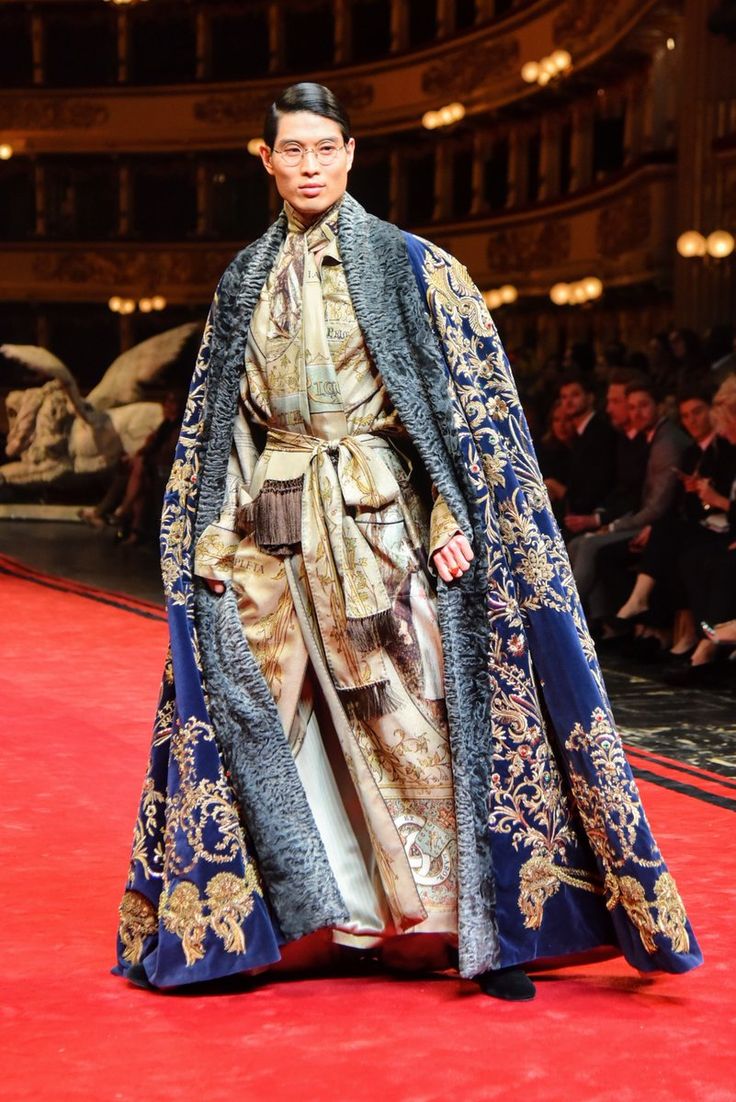a male model walks the runway in a stunning dolce & gabbana alta moda ensemble.  he wears a long, navy velvet cape with intricate gold embroidery and a grey fur trim. underneath, he sports a luxurious silk shirt and matching trousers in a beige and gold print.  the setting appears to be a grand opera house, adding to the dramatic and regal feel of the image. the model's posture and attire exude elegance and sophistication, highlighting the high fashion nature of the garment.  the red carpet adds to the opulent atmosphere.