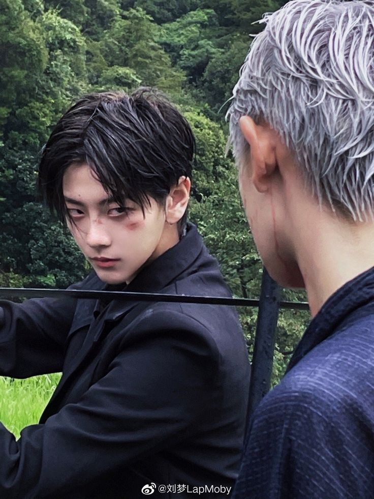 a photographic portrait featuring two young asian men, likely cosplaying, set against a verdant mountain backdrop. the man on the left, with dark hair and a bruise on his face, sports a serious expression and a black buttondown shirt. his gaze is directed intensely at the viewer. he appears to be holding something out of frame.  the other man, partially obscured, has striking silver hair and a visible scar on his ear.  he wears a bluegray shirt and is glancing back at his companion. the overcast lighting and lush greenery enhance the image's dramatic atmosphere.