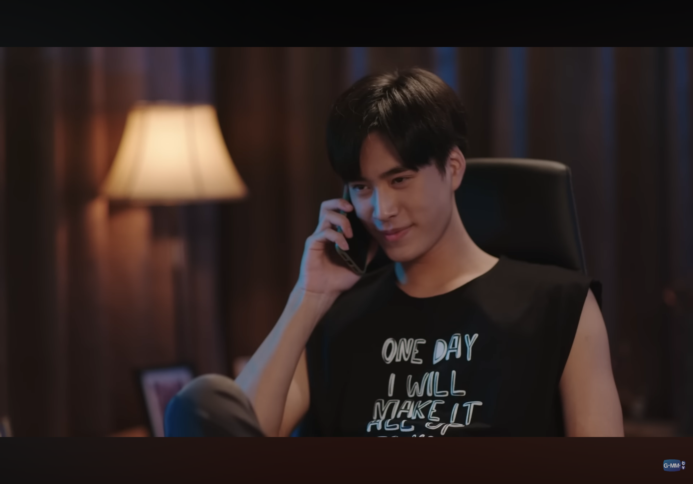 a young man with short black hair is sitting in a dimly lit room, talking on his phone and smiling. he's wearing a black sleeveless shirt with the words "one day i will make it all" printed on it in white. the overall mood of the image is casual and relaxed.