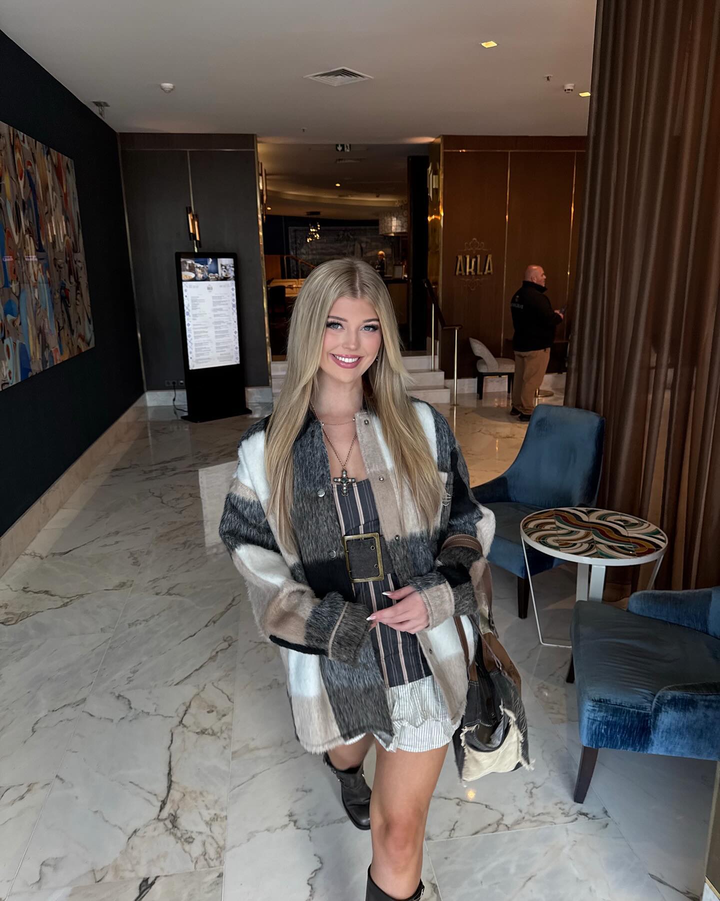 a young blonde woman is standing in a hotel lobby, smiling at the camera. she is wearing a plaid jacket, a short dress with a belt, and black boots. she is also carrying a handbag. the lobby has a marble floor and is decorated with artwork, chairs, and a table. there is a digital display and a person in the background.