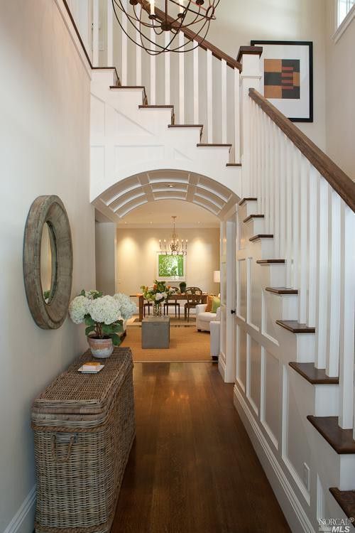 this image showcases the interior of a home, specifically a hallway with a staircase. the foyer area features a round mirror hanging on the wall above a wicker basket console table adorned with flowers. the design incorporates an archway leading into an open floor plan with a living room and dining room visible in the background. a chandelier hangs from the ceiling, illuminating the space. the staircase features a wooden railing and white balusters, leading to an upper level. the overall aesthetic is elegant and inviting, with a blend of classic and modern elements.