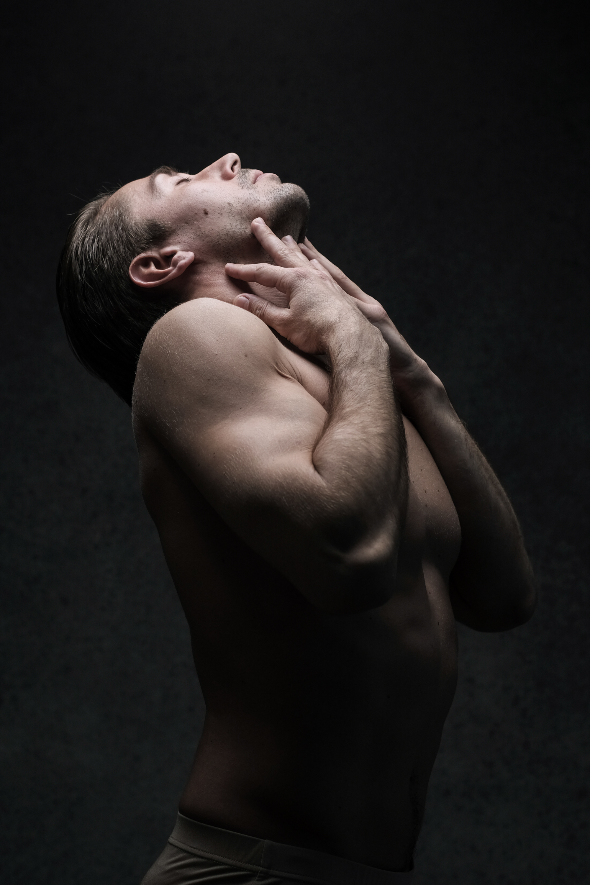 the image showcases a shirtless man against a dark, textured backdrop. he is positioned in profile, with his head tilted back and his eyes closed, appearing to be in a state