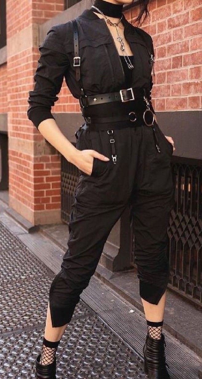 a woman in an allblack outfit poses on a city street. she is wearing black pants, boots, a choker, and a harness. she has her hair pulled back and looks edgy and cool. the background is a brick wall and a sidewalk.