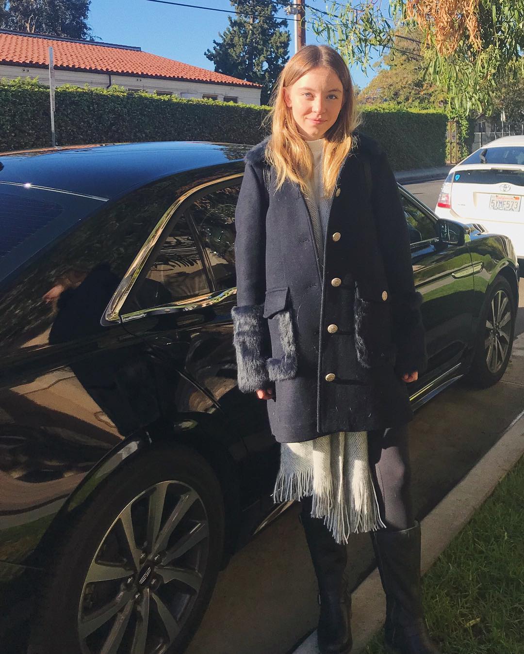 the image shows a young woman, possibly a teenager, standing next to a black luxury sedan. she's wearing a dark blue winter coat with fur trim on the cuffs and pockets, paired with black boots and a light grey scarf. the setting appears to be a residential street on a sunny day. the overall style is casual yet chic. the focus is on the girl, but the car and the background house are also visible.