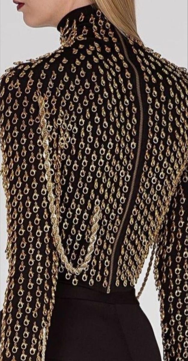 the image shows the back of a black highneck dress or top. the dress is decorated with numerous goldcolored loops or rings. a zipper runs vertically down the center of the back. a gold chain drapes down the side of the back.