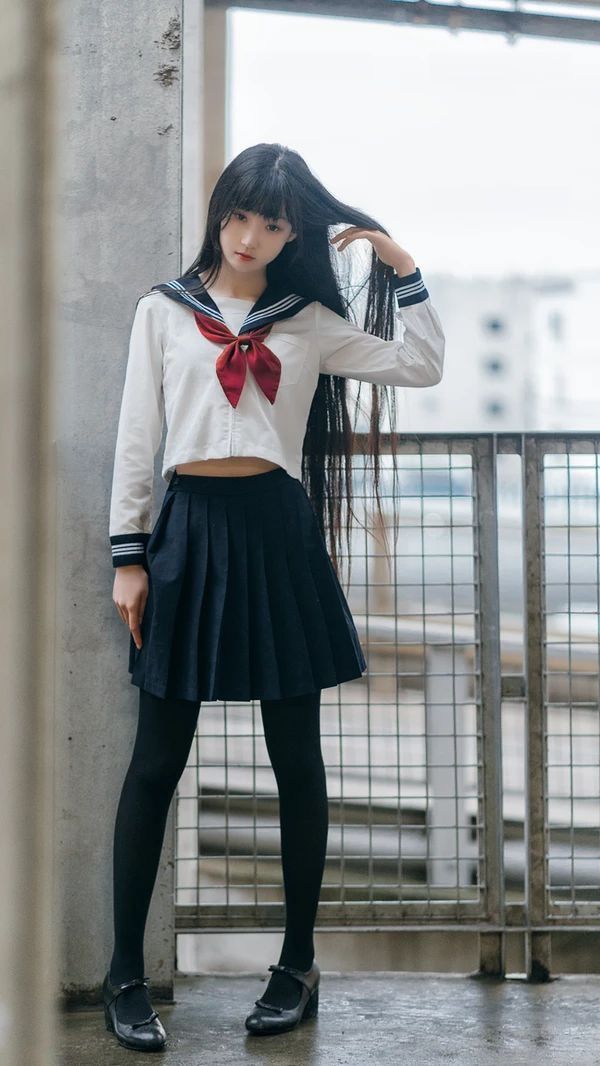a young asian woman is standing in front of a fence, wearing a school uniform with a sailor collar and red bow, black tights, and mary jane shoes. she has long black hair and is posing with one hand touching her hair. the background includes concrete and a building.