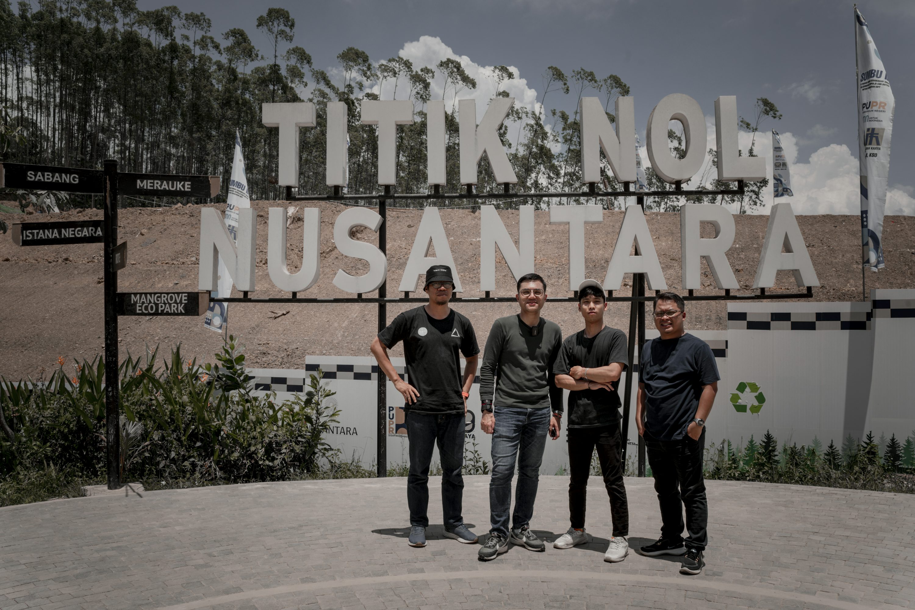a group of four young men stand proudly in front of the "titik nol nusantara" sign in indonesia. they are dressed casually and appear to be enjoying their time at this significant landmark. the background features lush greenery and a pathway, enhancing the scenic quality of the image.  directional signs indicating various locations including sabang, merauke, istana negara and mangrove eco park are visible on a post to the left. the overall vibe is one of adventure and exploration, showcasing a memorable travel experience.