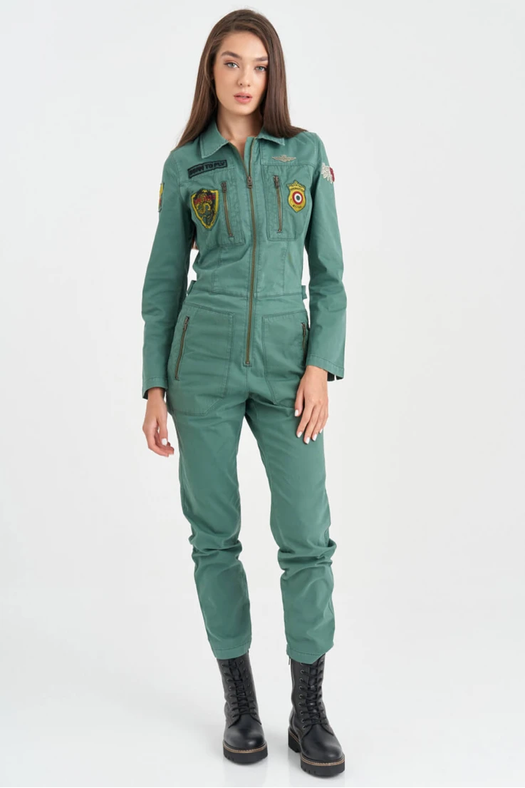 a young woman wearing a green jumpsuit with patches and black boots stands in a studio with a white background. she is posing with her hands at her side.