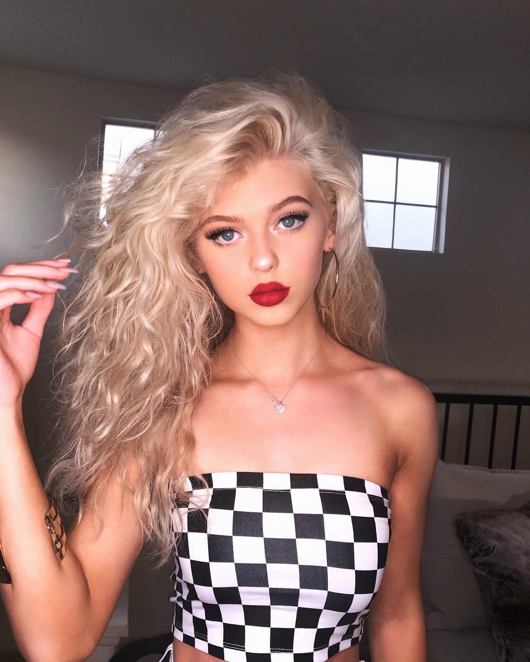 a young woman with long blonde, curly hair poses in front of two windows. she has blue eyes and is wearing red lipstick. she is wearing a black and white checkered strapless top. she is also wearing hoop earrings, a necklace, and an arm bracelet. the background appears to be a bedroom.