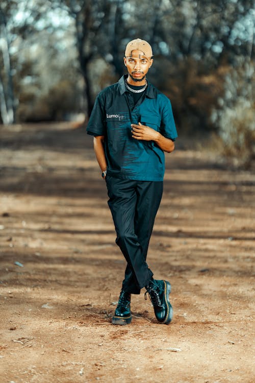 a young man stands on a dirt path in a forest. he is wearing a teal shirt, black pants, a beige cap, and black boots.  he has a serious expression on his face and is looking directly at the camera. the background is blurred but shows a natural setting with trees. the overall style is streetwear.