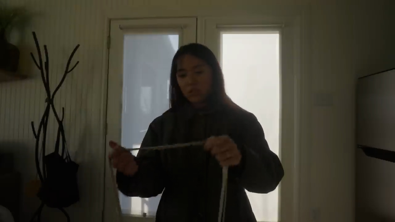 a young woman stands in a room with natural light streaming in from a window behind her. she is holding a measuring tape in her hands, seemingly preparing to measure something. she is wearing a dark jacket over a hooded sweatshirt. the overall ambiance is one of quiet focus and creative energy.