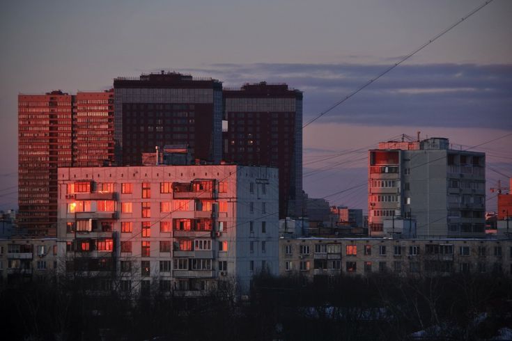 a stunning cityscape at sunset.  the image features several multistory apartment buildings, some taller than others.  the setting sun casts a warm, reddishorange glow on the windows of a central building, creating a striking contrast against the cooler tones of the other buildings and the darkening sky.  power lines crisscross the scene, adding to the urban feel.  silhouetted trees in the foreground partially obscure the base of the buildings, adding depth to the composition.  the overall mood is serene yet urban.