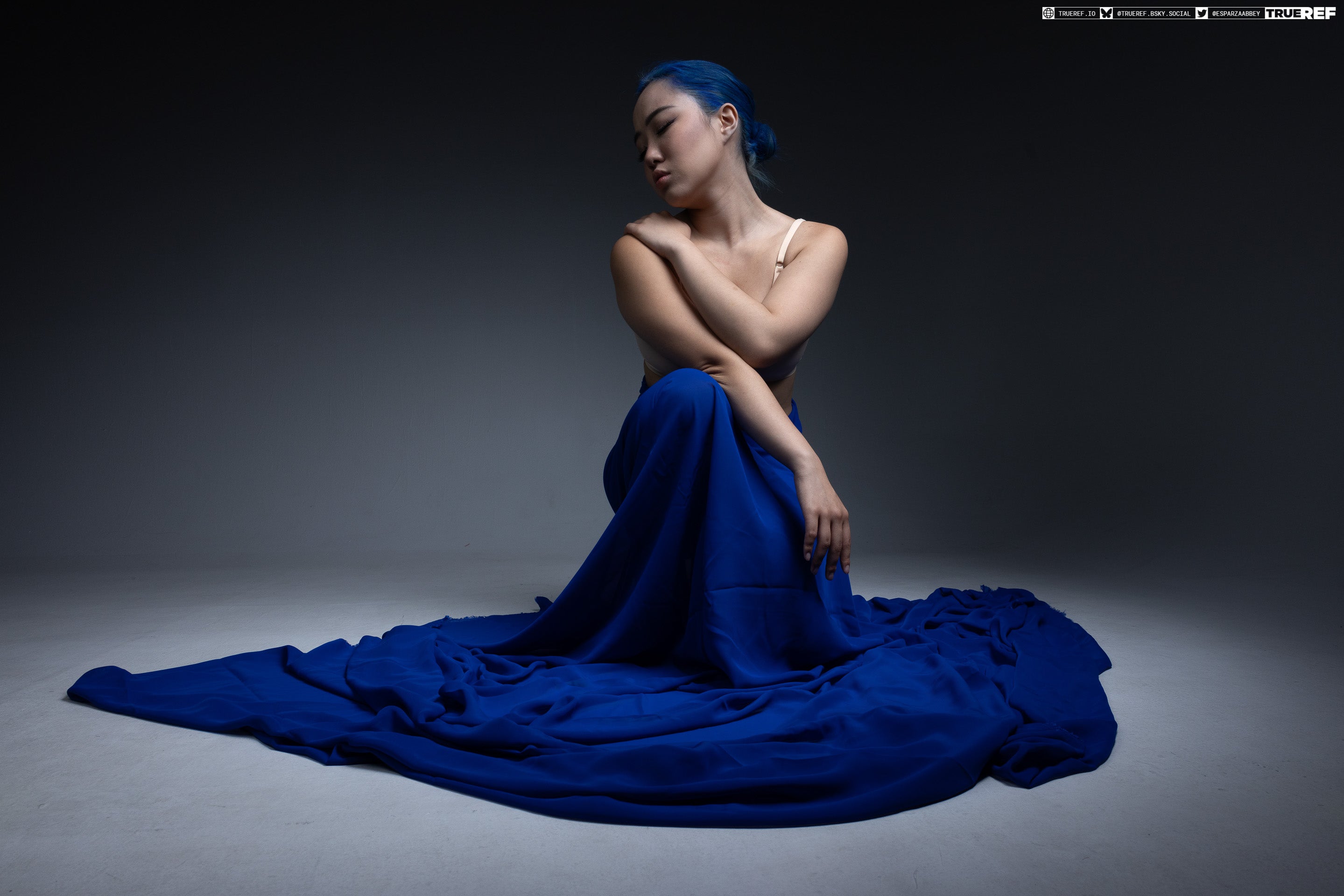 a studio shot of a woman with blue hair wearing a bra, sitting down with arms crossed and eyes closed. she is draped in a large blue fabric.