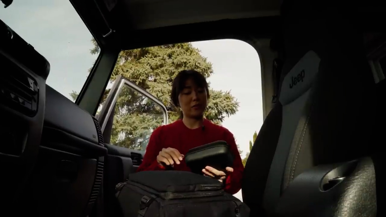 a young woman is packing her camera gear into a black backpack while sitting in the driver's seat of a jeep.  the door is open, and we can see a glimpse of greenery outside the vehicle. the scene is bathed in natural light, hinting at an overcast day. the perspective suggests the viewer is also inside the car, possibly about to embark on an adventure with the woman.