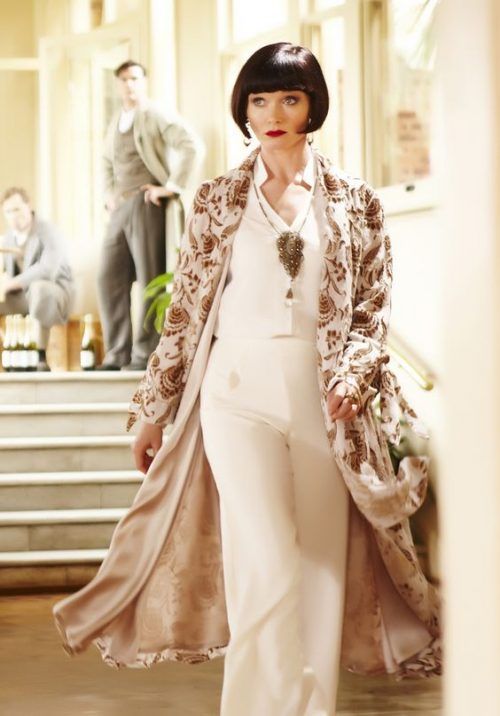 a woman in a long white dress with brown floral print on it, standing on a staircase and looking down. she has a bob haircut and red lips.