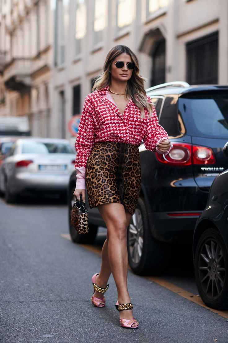 a stylish woman in a red and white gingham shirt and leopard print skirt walks down a city street. she is wearing pink high heels with gold chain details, sunglasses and carrying a leopard print handbag.  several cars are parked along the street in the background. the overall style is trendy and chic.