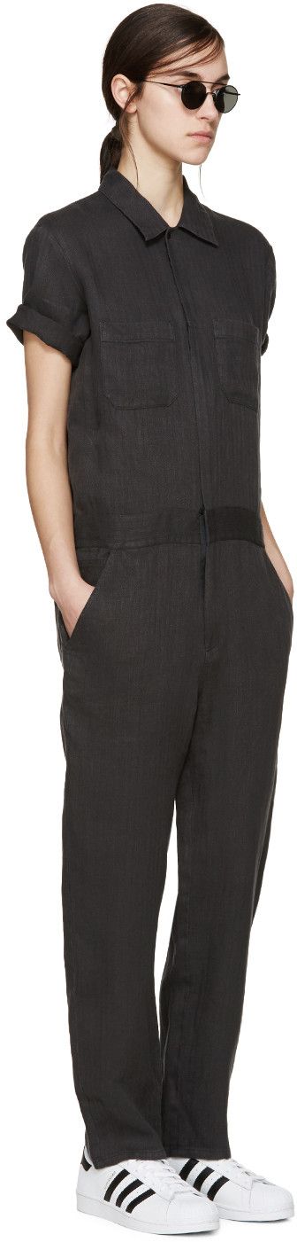 a woman poses in a black shortsleeved linen jumpsuit, wearing white sneakers and dark sunglasses. the jumpsuit has a collar and two pockets on the chest.