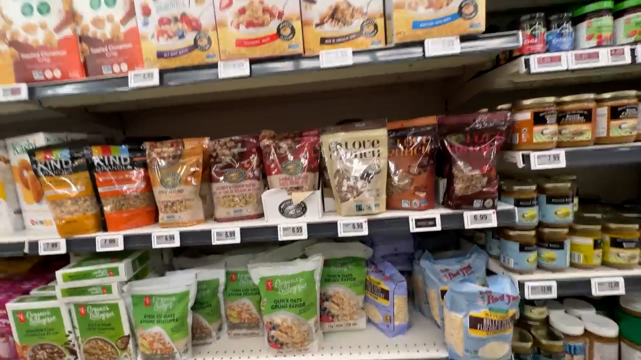 a view across multiple shelves in the breakfast food aisle of a grocery store. the shelves are stocked with various brands and flavors of cereal, granola, and other breakfast items. the packaging is colorful and eyecatching. the lighting is bright and fluorescent.