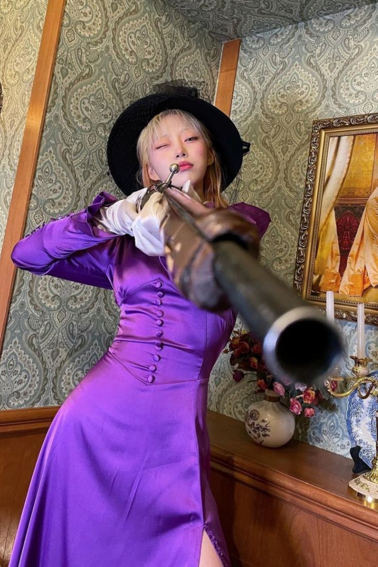 a woman wearing a purple dress and a black hat is holding a gun while looking directly at the camera. she is standing in a vintage setting with patterned wallpaper and gold decor. she has a serious expression