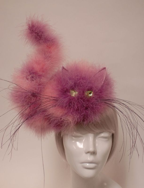 a mannequin head wearing a novelty catshaped hat made of pink and purple feathers. the hat has a curved tail, whiskers, and jeweled eyes.